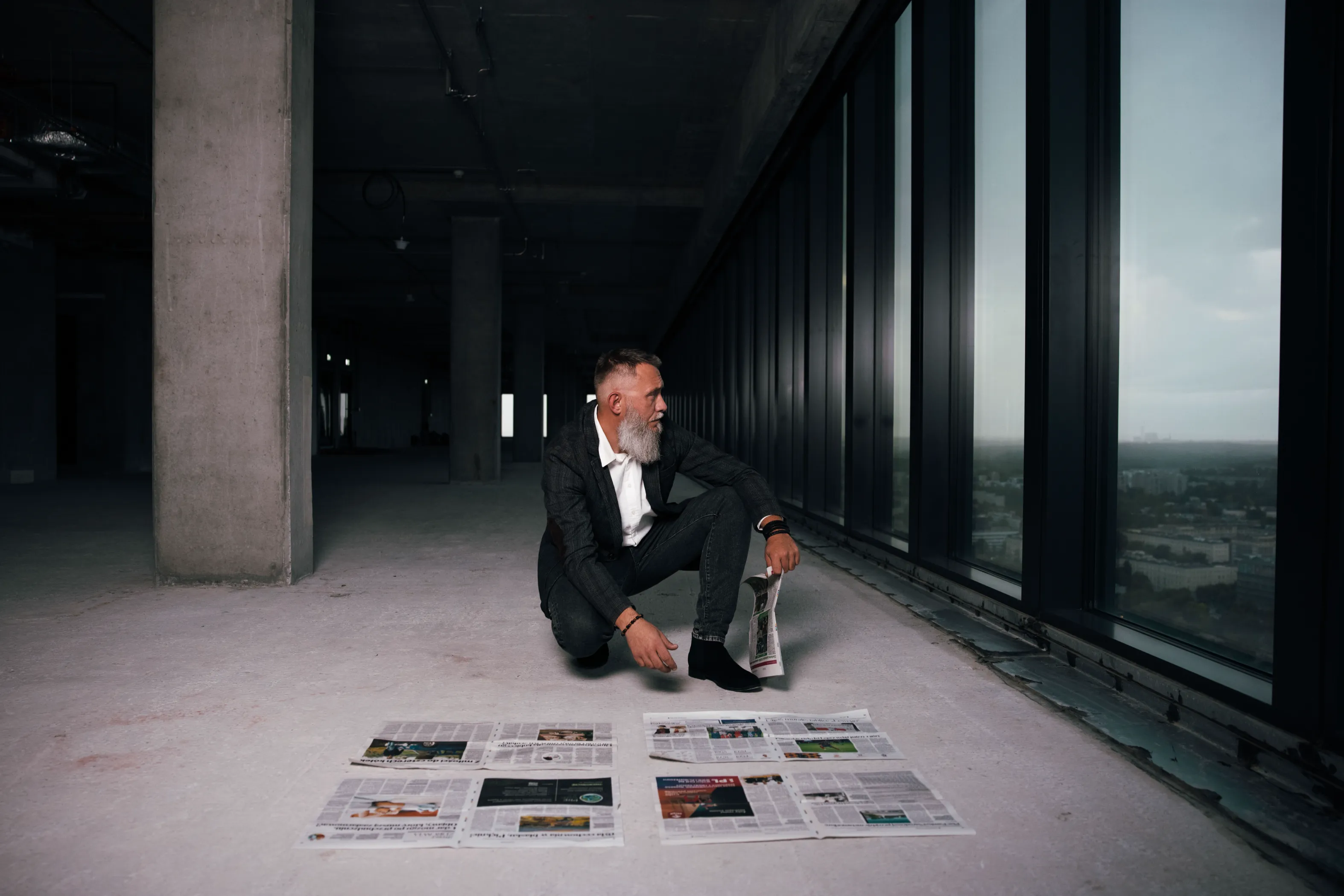 Retrato corporativo en rascacielos de Santo Domingo Hombre de barba cana en traje negro en cuclillas frente a ventanal con vistas de Santo Domingo, luz natural difusa, retrato corporativo profesional