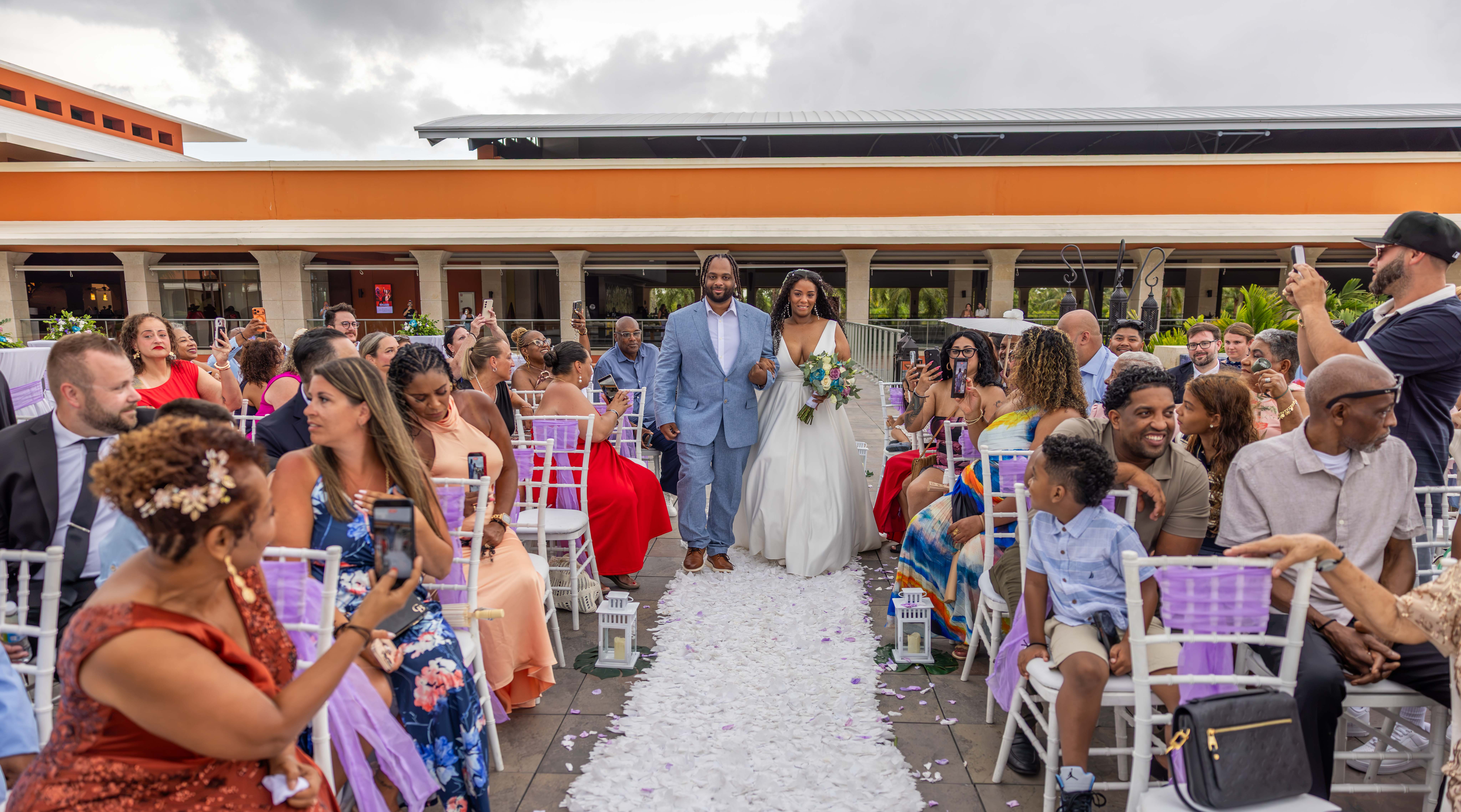 Novios caminan bajo toldo naranja en boda playera Novios caminan por el pasillo en boda de playa en Santo Domingo bajo toldo naranja, rodeados de familia celebrando