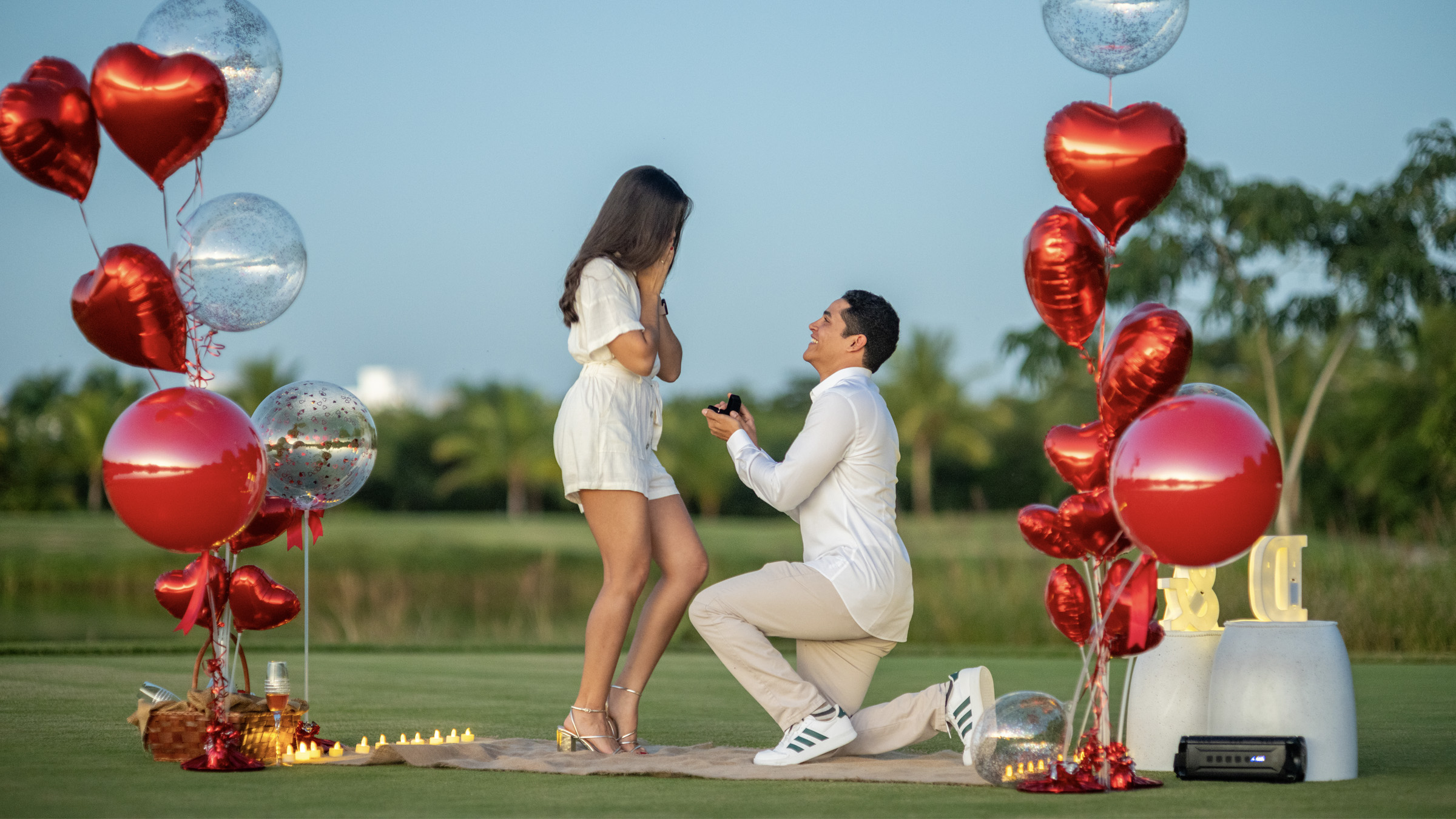 Marriage proposal beneath red balloons Engagement proposal in open field in Santo Domingo: man on one knee before smiling woman, surrounded by red and clear balloons, cloudless blue sky and palms in background