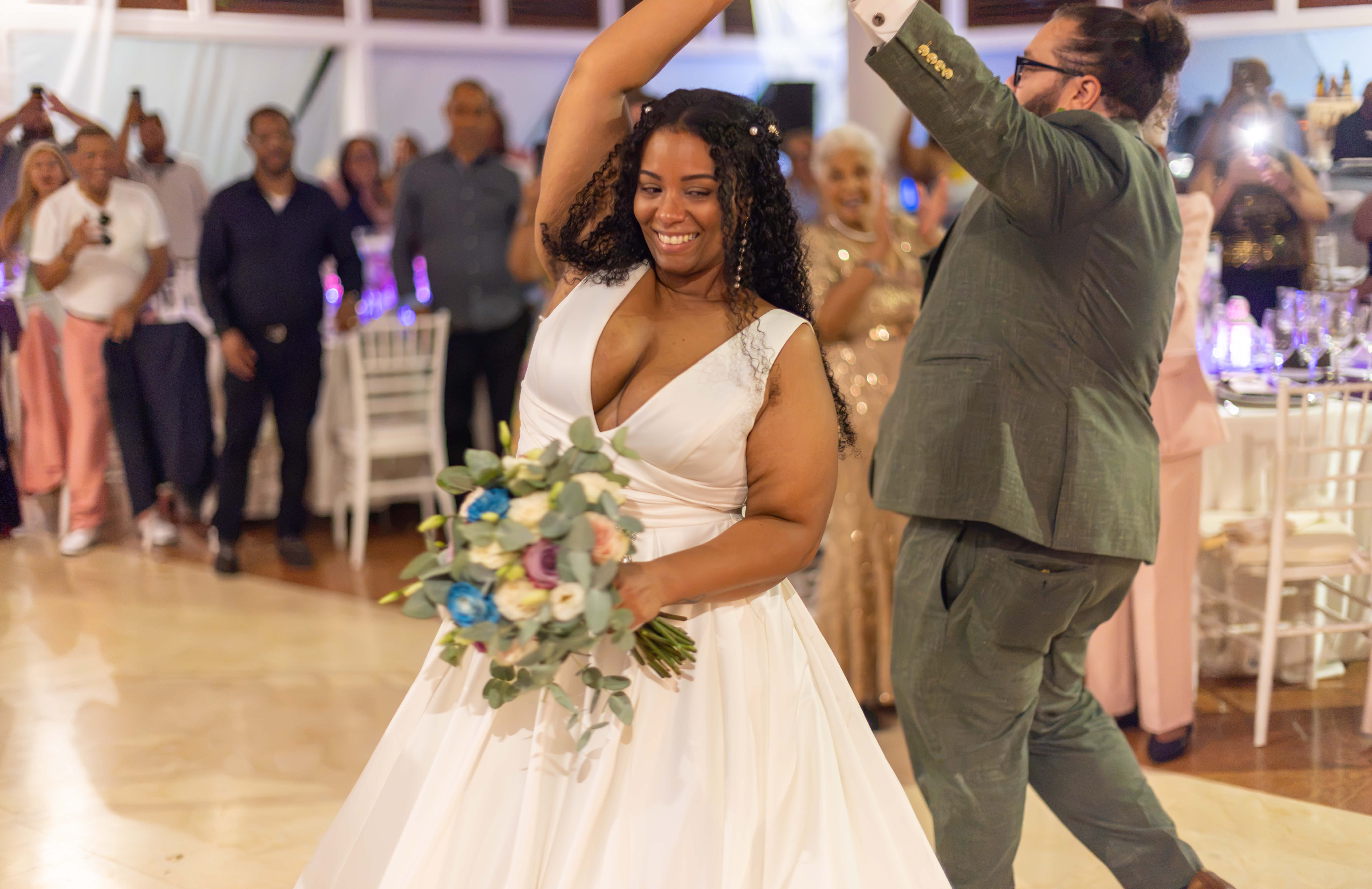 Primer baile radiante de novia y novio Novia y novio en primer baile durante boda en Santo Domingo: ella sonríe radiante con ramo azul, él la levanta en piso de madera, invitados celebran alrededor