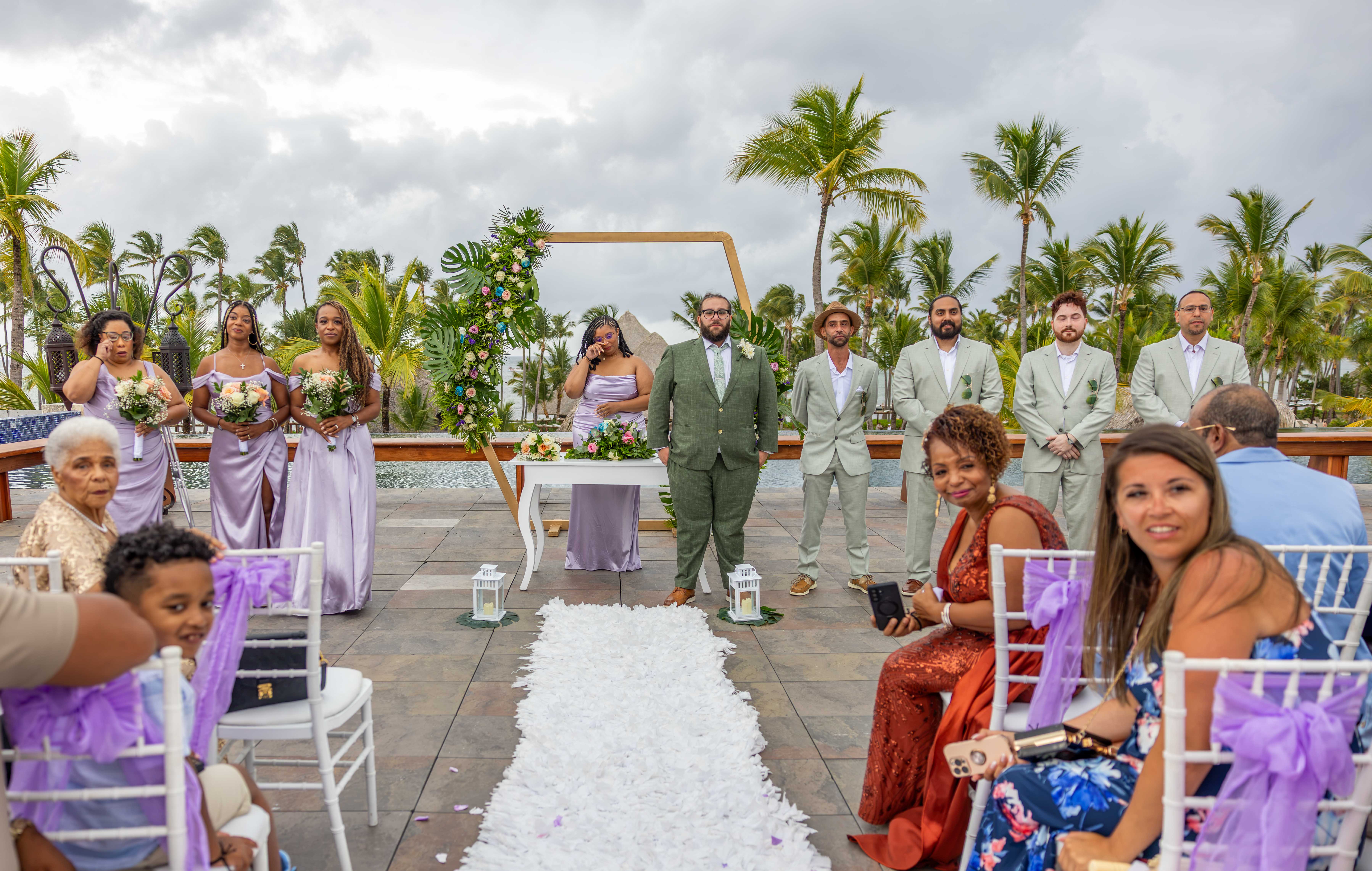 Caribbean seaside wedding ceremony under wooden arch Beach wedding ceremony with bride, groom and wedding party under wooden arch, white petal aisle with palm trees in background