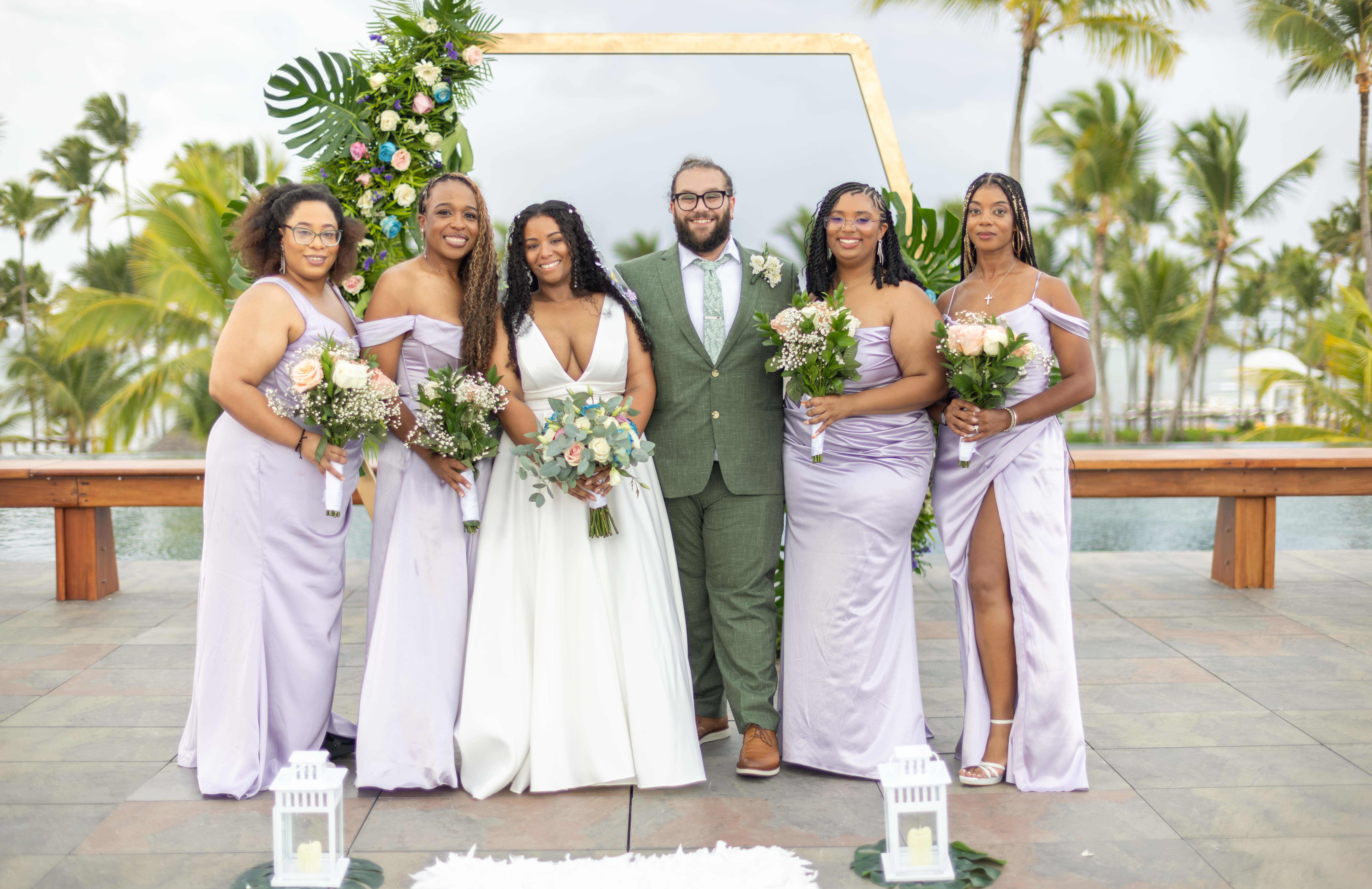Retrato grupal de bodas playera Grupo de novia, novio y cinco damas de honor en vestidos lavanda y musgo frente a marco dorado con palmeras de fondo. Fotógrafo de bodas en Santo Domingo.