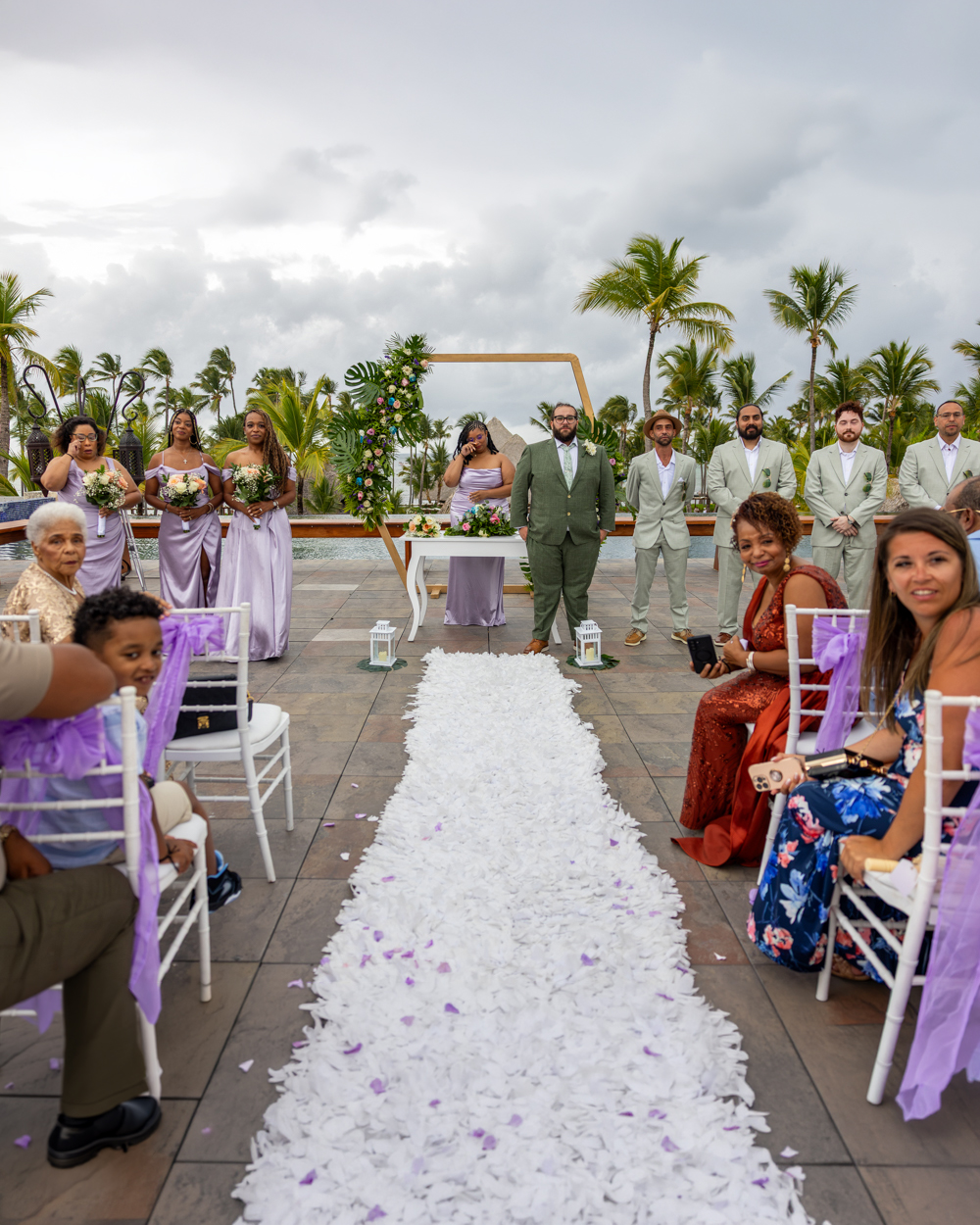 Ceremonia nupcial bajo palmeras caribeñas Ceremonia de boda tropical en Punta Cana con arco de madera, pareja intercambiando votos, palmeras de fondo y pétalos púrpura sobre arena blanca