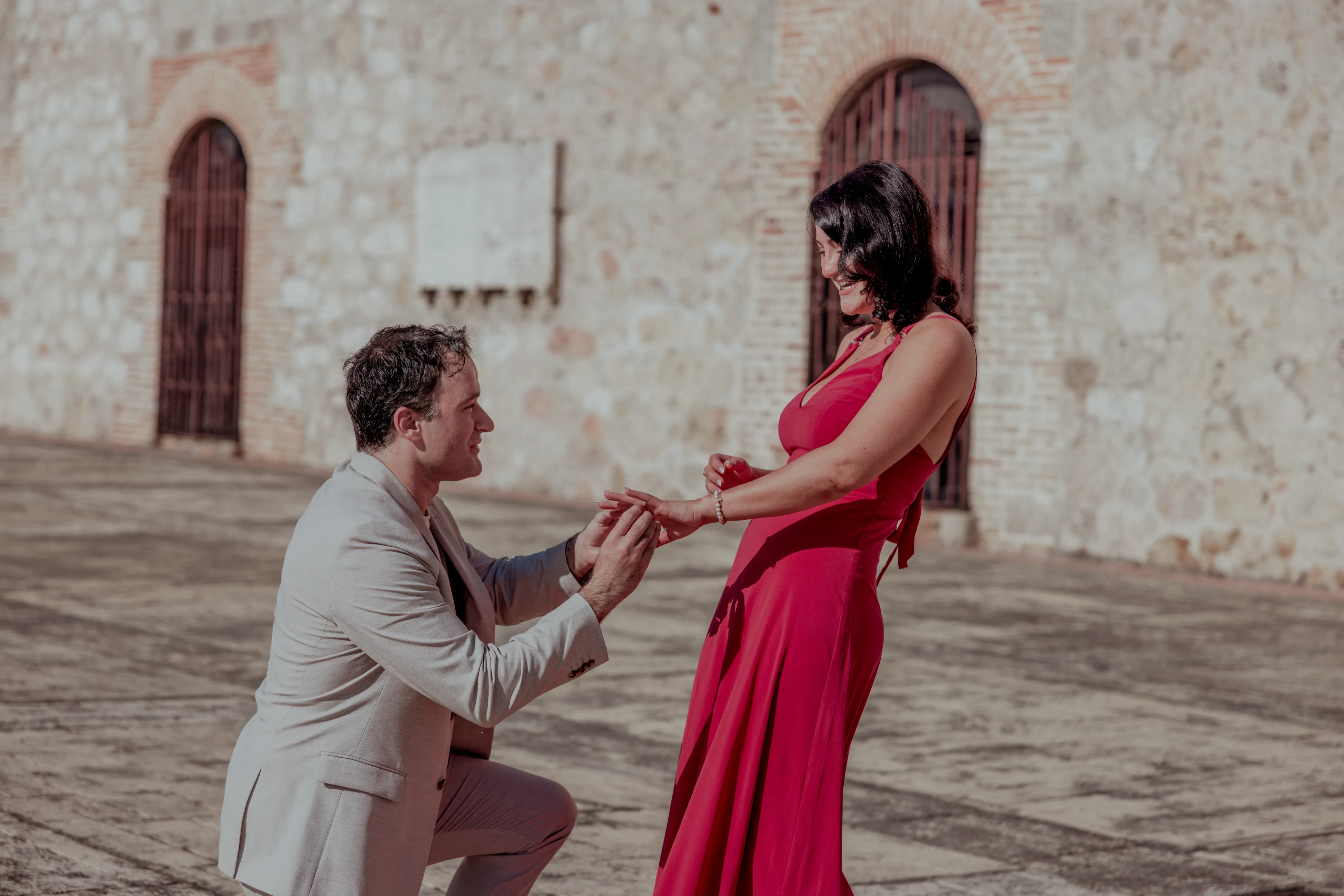 Propuesta matrimonial en la Zona Colonial Hombre en traje beige se arrodilla con anillo frente a mujer en vestido fucsia en plaza colonial de Santo Domingo